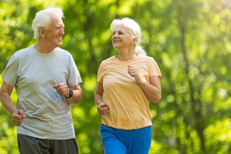 Idade mental pode ser diferente da cronológica. Um casal de idosos correndo no parque ao céu aberto.
