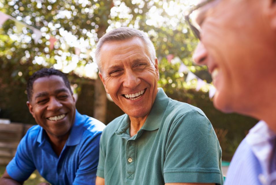 Um grupo de amigos, três homens idosos, em uma área aberta, sorrindo. Imagem para ilustrar a matéria sobre prevenção do Alzheimer.